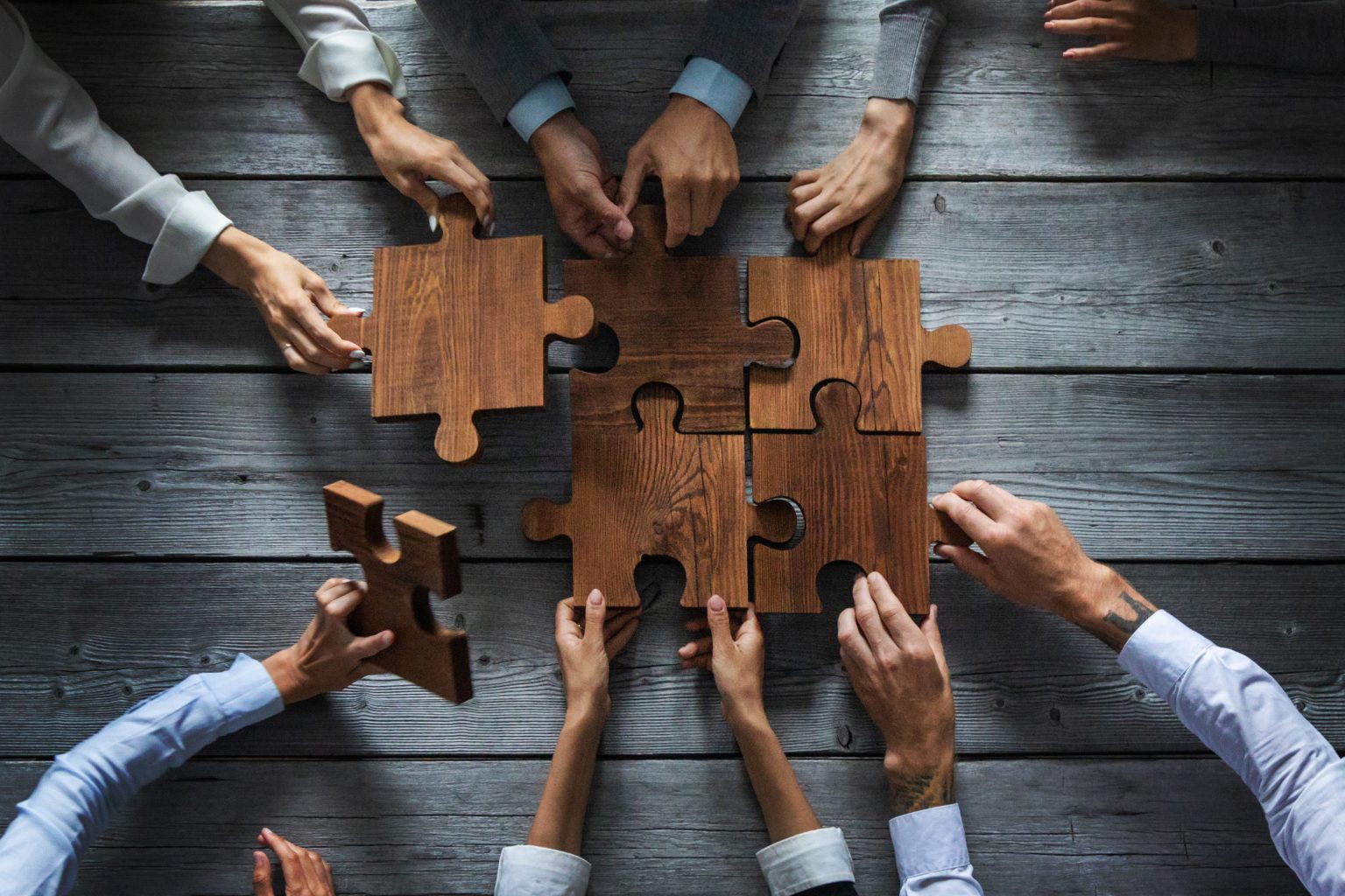 Sontro - Zaposlitev 1 Sontro - business people team sitting around meeting table assembling wooden jigsaw puzzle pieces unity cooperation ideas concept